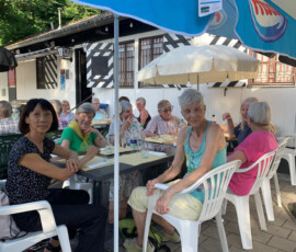 Group of elderly people sitting at outdoor cafe tables under umbrellas, enjoying a meal together.