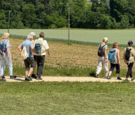 Group of hikers with backpacks walking along a dirt path through a rural field, with trees and hills in the distance.