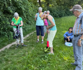 A group of older adults on a grassy path in a park, some with backpacks, chatting and resting.