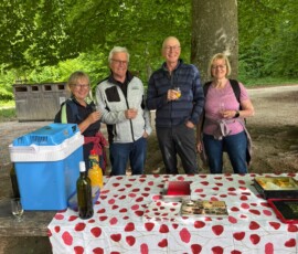 #image_title Four friends stand together outdoors under a leafy tree, holding drinks and smiling at the camera beside a picnic table with a red-rose tablecloth and a blue cooler.