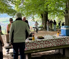 #image_title Group of people chatting around a long table in a park under green trees, with a heart-patterned tablecloth and drinks nearby.