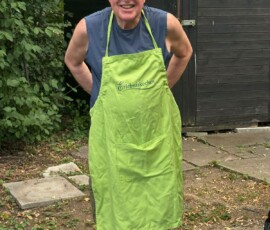 Smiling older man in a bright lime apron standing outdoors in front of a dark wooden shed; blue sleeveless shirt visible underneath.