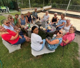 A group of about 14 mature adults sitting in a circle around a table outdoors, sharing food and drinks on a sunny lawn behind a sports court.