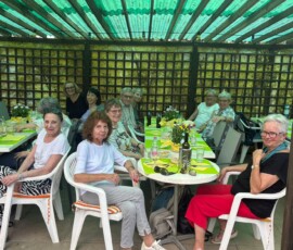 Group of seniors seated around a long outdoor dining table with green placemats under a pergola, smiling and chatting.