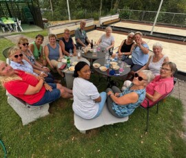 Group of seniors seated around an outdoor table enjoying snacks and drinks in a sunny yard setting.