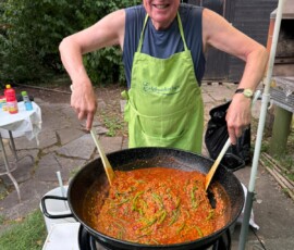 Older man with a lime-green apron labeled 'Erlebniskocken' smiling as he stirs a large pot of red chili outdoors on a barbecue burner under a yellow-striped umbrella.