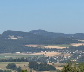 Wide landscape of rolling hills and patchwork fields with a small village in the valley, framed by trees in the foreground under a clear blue sky.