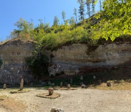 Steep rock cliff with bushes and trees above, under a bright blue sky, and numerous upright stone markers arranged on the dirt ground in front of the rock face.